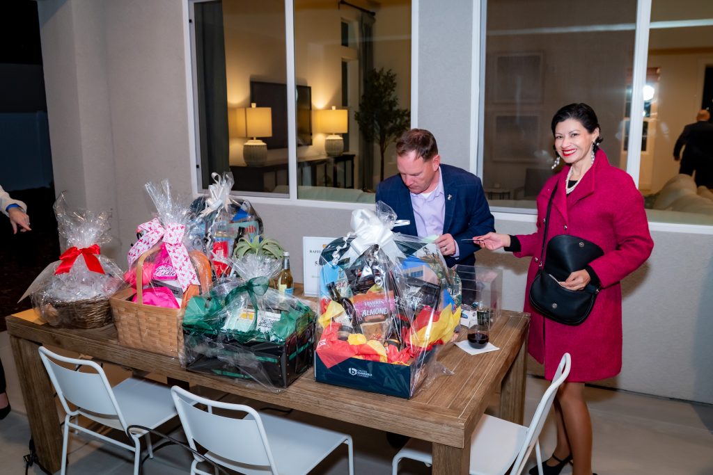 A man sits at a wooden table with several colorful gift baskets, while a woman in a bright pink coat stands nearby, smiling and holding a black purse. They appear to be at an indoor event or gathering.