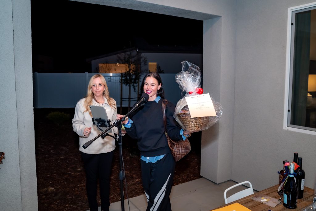 Two women stand under a covered patio at night. One holds a microphone and a gift basket wrapped in cellophane, smiling, while the other holds a clipboard and looks at the camera. A table with wine bottles is nearby.