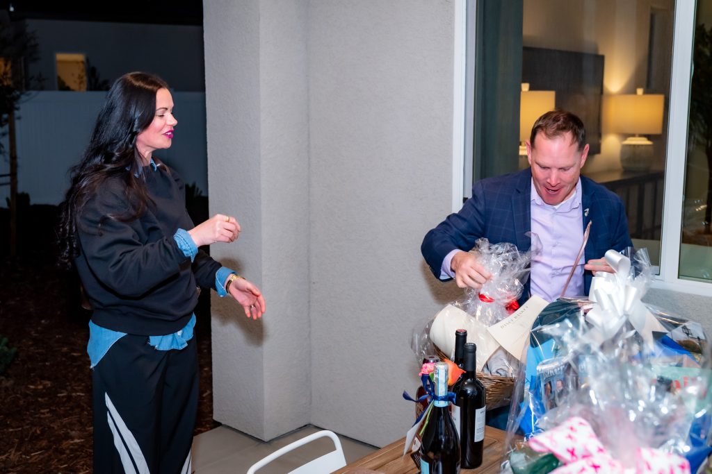 A woman stands smiling while a man in a suit examines a gift basket on a table filled with bottles and wrapped presents, indoors with bright lighting.