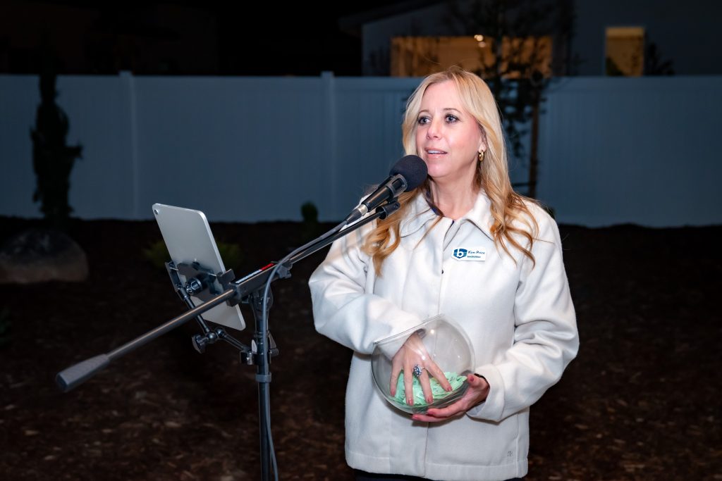 A woman stands at a microphone outdoors at night, holding a glass bowl with green slips of paper. She wears a light-colored jacket and looks slightly to the side. A tablet is mounted on the microphone stand.