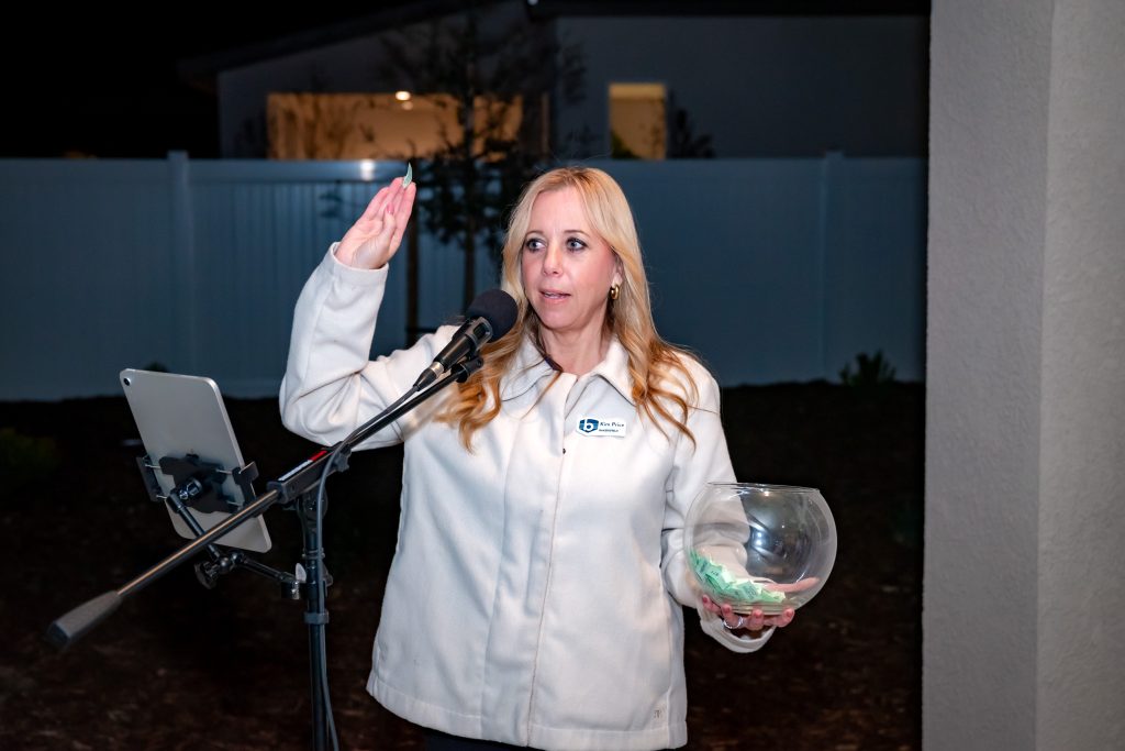 A woman stands in front of a microphone at night, raising her right hand as if taking an oath. She is holding a clear bowl with tickets or slips in her left hand and wearing a white jacket. A house and white fence are in the background.