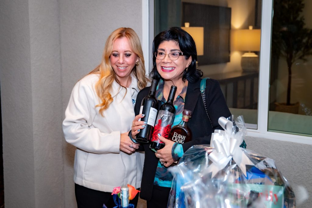 Two women smile together indoors; one woman holds bottles of wine and whiskey, while the other stands beside her. A gift basket wrapped in cellophane with a white ribbon is visible in the foreground.