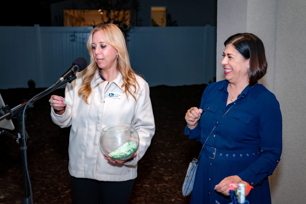 Two women stand together; one draws a slip of paper from a bowl in front of a microphone, while the other woman smiles. They appear to be participating in a raffle or drawing at an outdoor event.