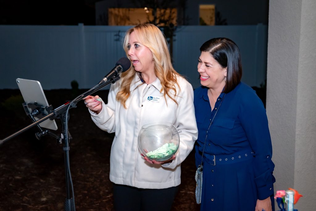 Two women stand beside a microphone outdoors at night. The woman on the left holds a glass bowl with green papers and speaks into the mic, while the woman on the right smiles and looks at her. A tablet is mounted in front of them.