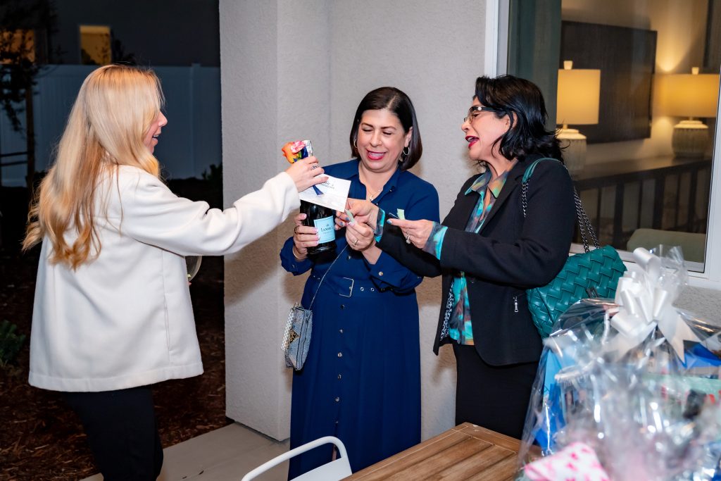 Three women stand together outside near a table with a gift basket. One woman hands a bottle and card to another, while the third woman looks on, smiling and holding a piece of paper. They appear to be at a social gathering.