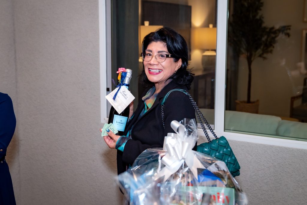 A woman with black hair and glasses smiles while holding a blue bottle with a tag and a large gift basket wrapped with a white bow. She is indoors, wearing a dark jacket and has a green purse over her shoulder.