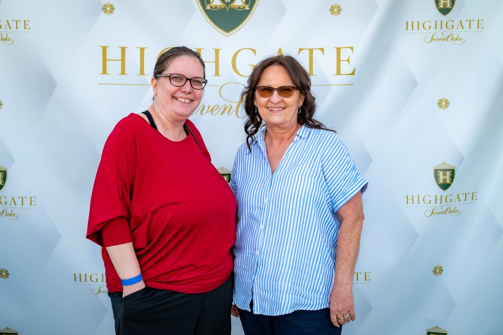 Two women smiling and posing together in front of a step-and-repeat banner with a “HighGate” logo and gold text. Both wear glasses; one is in a red top, the other in a blue-and-white striped shirt.