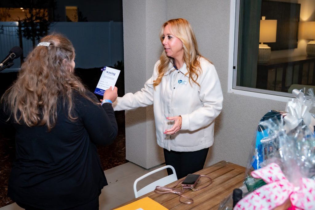 Two women are standing indoors; one hands a certificate to the other. The woman receiving the certificate wears a white jacket and gestures with her left hand. There are gift baskets and a phone on the table nearby.