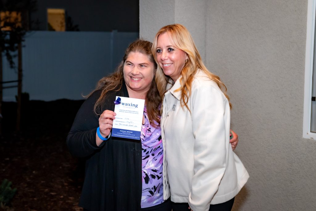 Two women smile and pose together outside at night. The woman on the left holds a waxing gift certificate, while the woman on the right, dressed in a white jacket, stands close beside her.