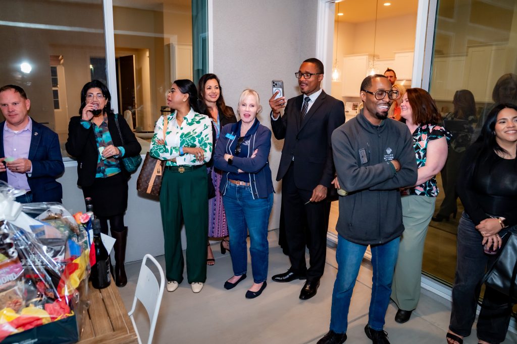 A group of people stand together indoors, smiling and laughing as they watch something off-camera. Some hold drinks and a man is taking a photo with his phone. A gift basket sits on a table in the foreground.
