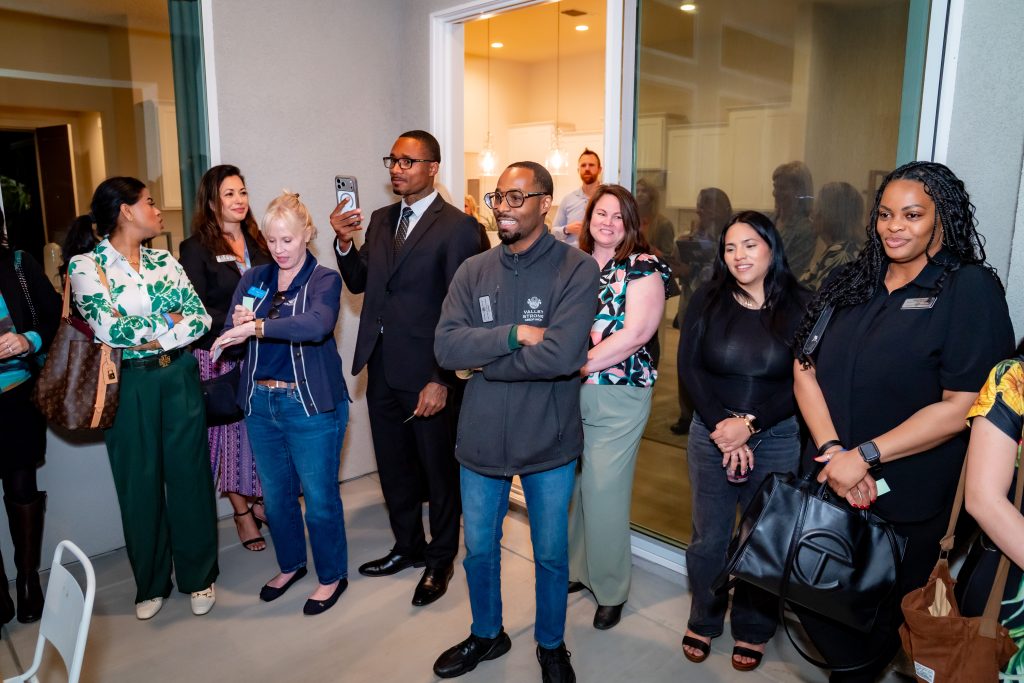 A diverse group of people stand together indoors, smiling and watching something off camera. Some are holding phones, while others have their arms crossed or hands clasped. It appears to be a casual, social gathering.