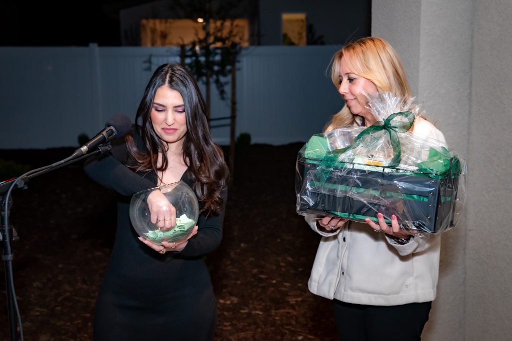 Two women stand outside; one draws a paper from a glass bowl near a microphone, while the other holds a wrapped gift basket and smiles at her.