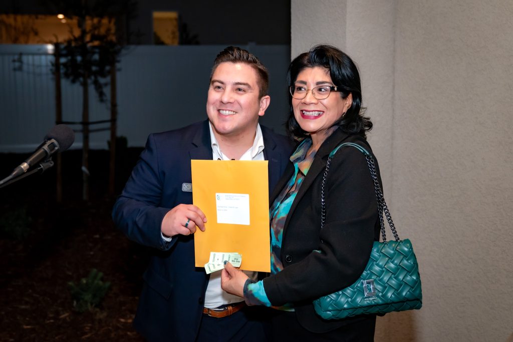 Two people smiling and posing together indoors; the man is holding a large yellow envelope and some keys, while the woman stands beside him with a teal handbag.