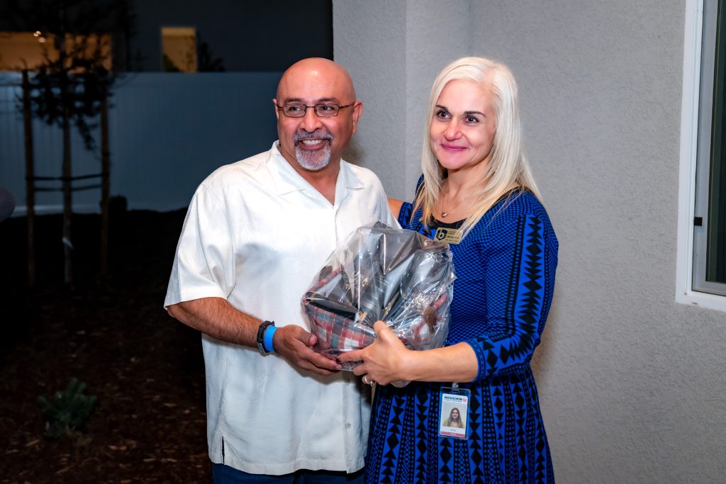 A smiling man in a white shirt stands next to a woman in a blue dress holding a wrapped gift basket. They appear to be at an indoor event near a window, with a dark outdoor area in the background.