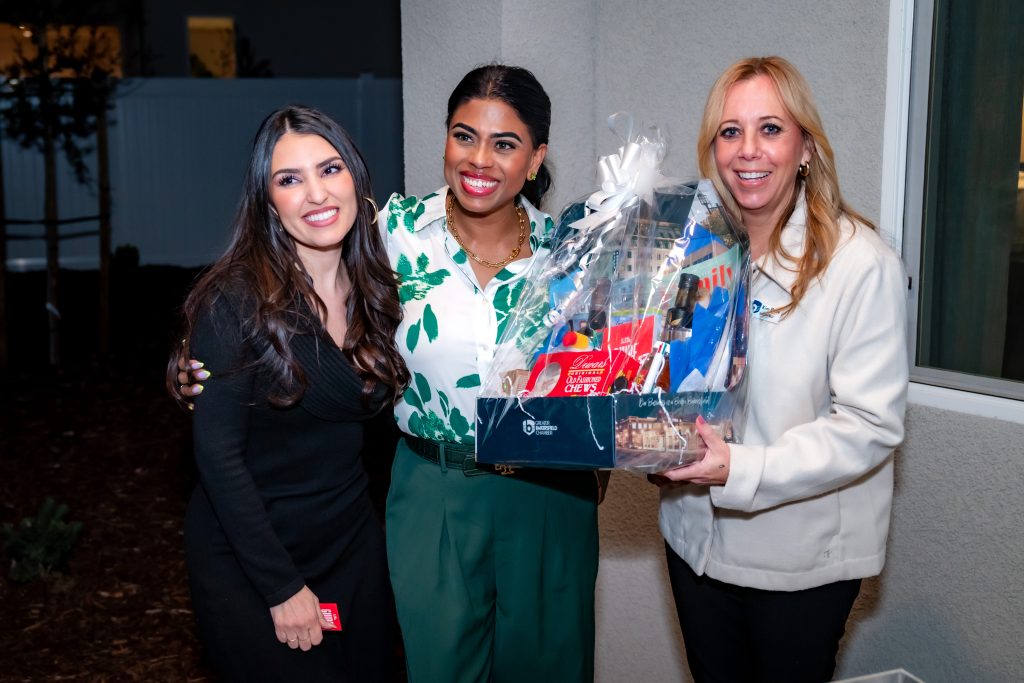Three women stand together smiling, one holding a large gift basket wrapped in clear plastic. They are dressed in semi-formal clothing and are posing indoors near a light-colored wall, appearing happy and celebratory.