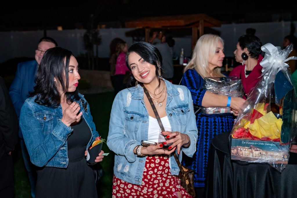 Two women stand by a table at an outdoor event. Both wear denim jackets; one smiles at the camera while holding a phone, the other looks at her. People and a gift basket are visible in the background.