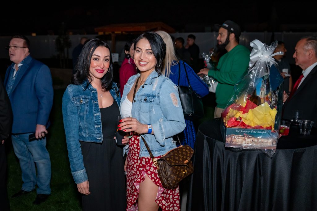 Two women smiling and posing together at an outdoor event, both wearing denim jackets. People are mingling in the background, and a gift basket sits on a nearby table. The atmosphere is festive and lively.