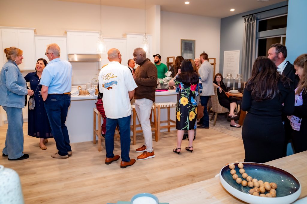 A group of people mingle, chat, and laugh in a brightly lit kitchen and living room during a casual indoor gathering. Some stand in conversation clusters, while others sit on chairs or stools.
