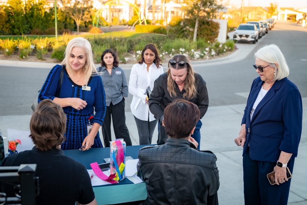 A group of people, mostly women, gather around a table outdoors on a sunny day. Some are standing, talking, or interacting with items on the table, while two people sit with their backs to the camera. Cars are parked along the street.