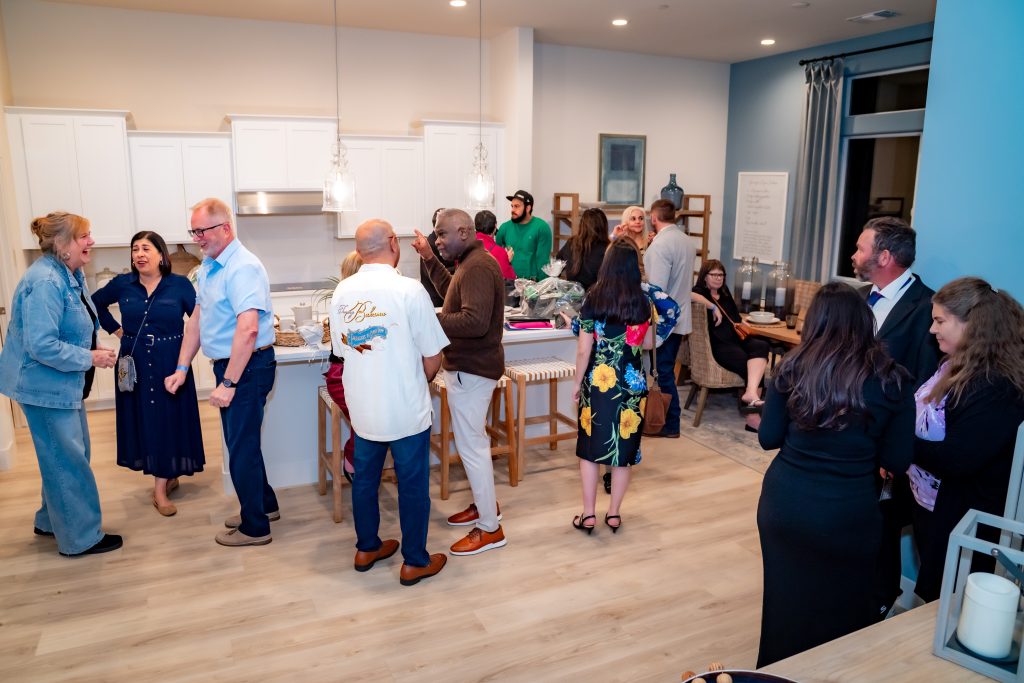 A group of adults socializing at a gathering in a modern kitchen and living area. Some are chatting in small groups, while others stand near the kitchen island. The atmosphere appears friendly and lively.