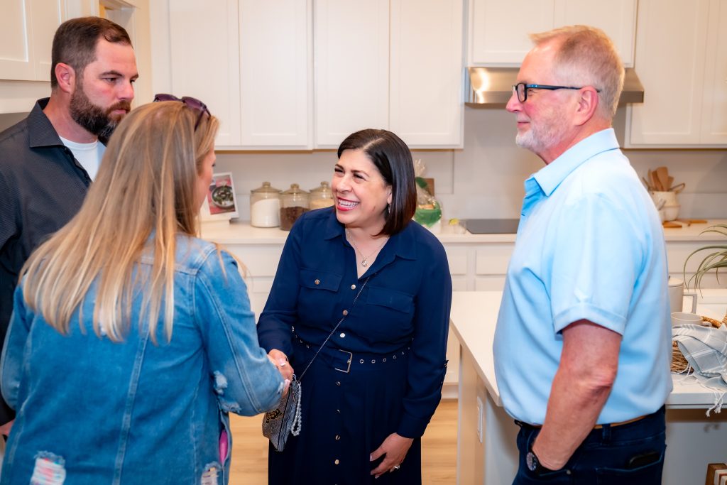 Four adults stand in a bright kitchen, two women shaking hands and smiling, while two men watch and chat nearby. Everyone appears engaged and friendly.