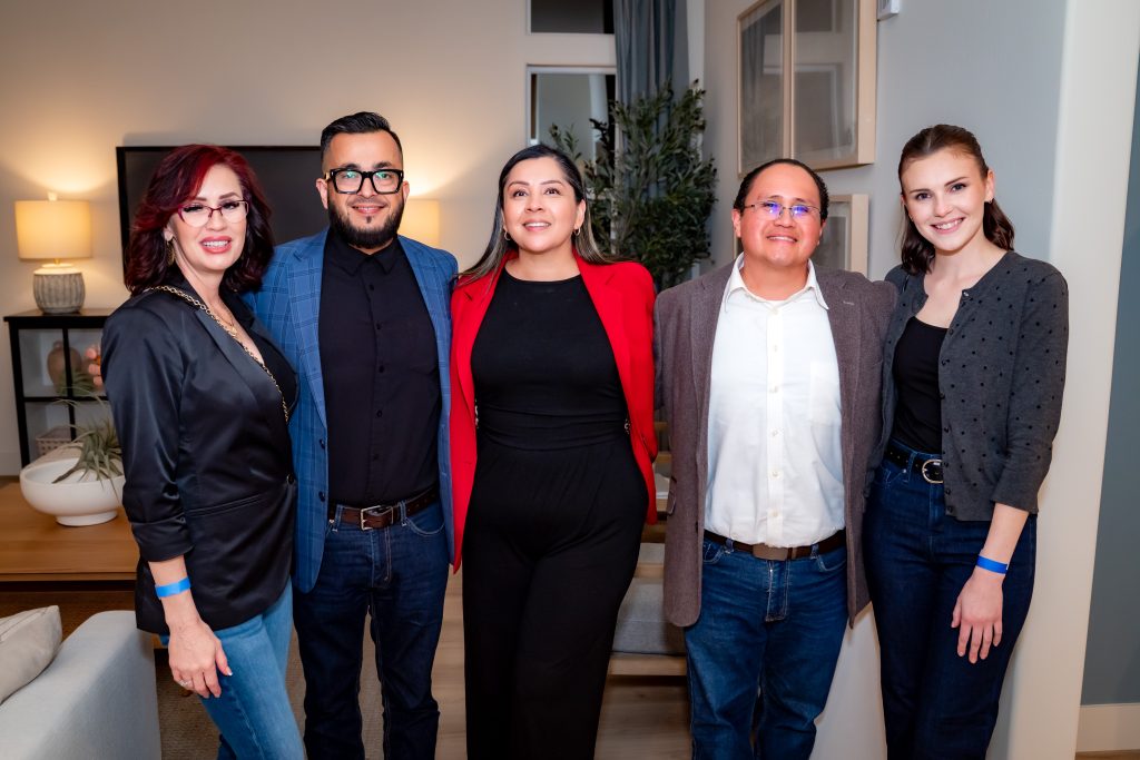 Five people stand together indoors, smiling at the camera. They are dressed in semi-formal attire, with a well-lit modern living room setting in the background.