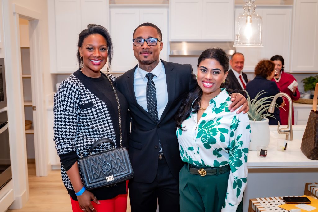 Three people pose and smile together in a bright, modern kitchen during a gathering. The man in the center wears a suit, with women on either side dressed stylishly. Other guests are chatting in the background.