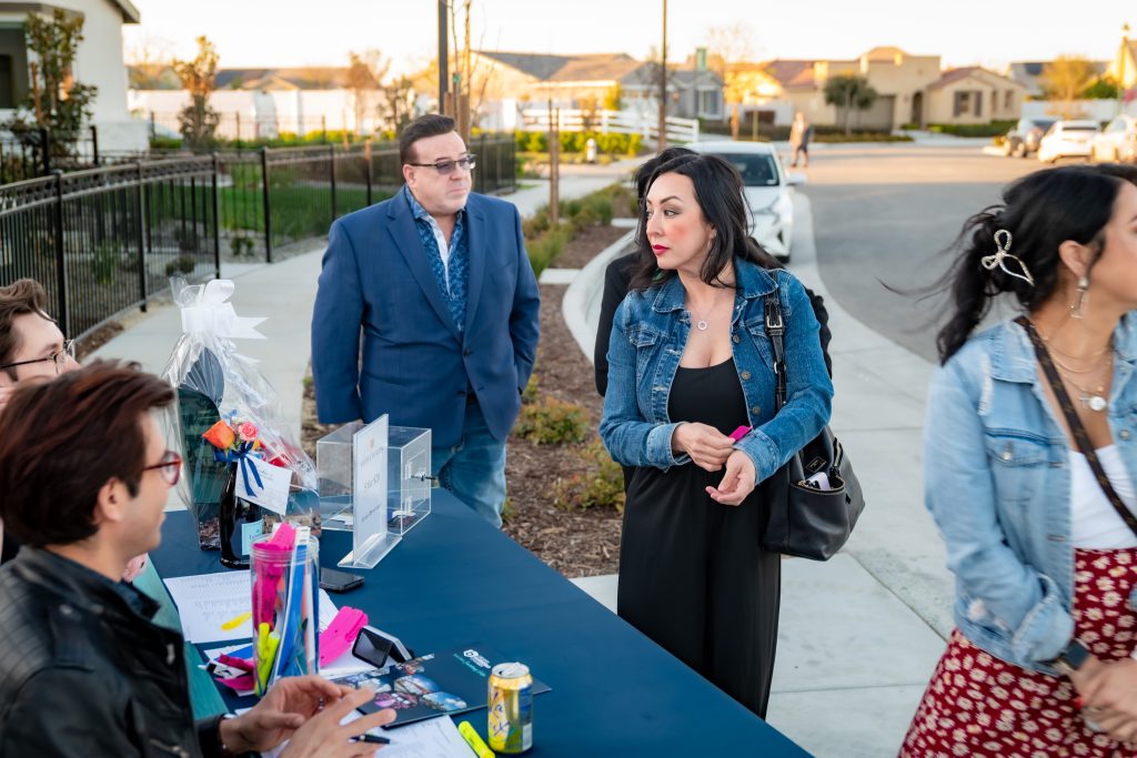 A woman in a denim jacket stands near a check-in table outdoors, talking to seated staff. A man in a blue suit stands nearby, and another woman in a patterned skirt is partly visible. Houses and cars are in the background.
