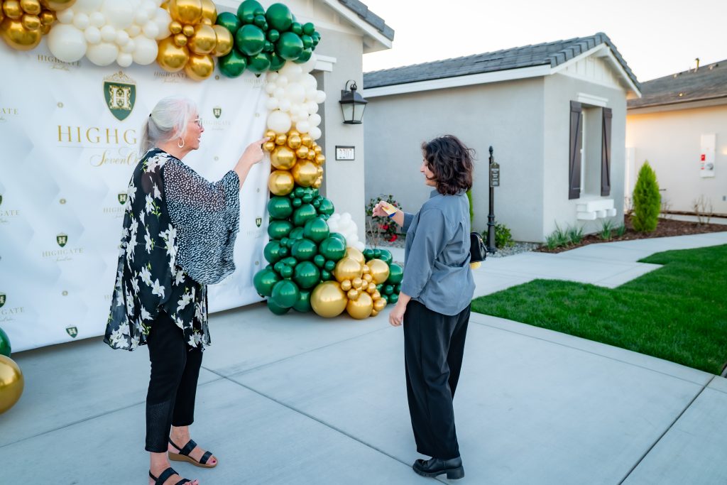 Two women stand outside a house near a decorative balloon arch in green, gold, and white, in front of a backdrop that says “Highgate.” One woman gestures toward the arch while the other listens.