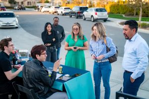 A group of people stand and sit around a blue table set up on a suburban street, talking and interacting in a casual outdoor setting with cars and houses visible in the background.