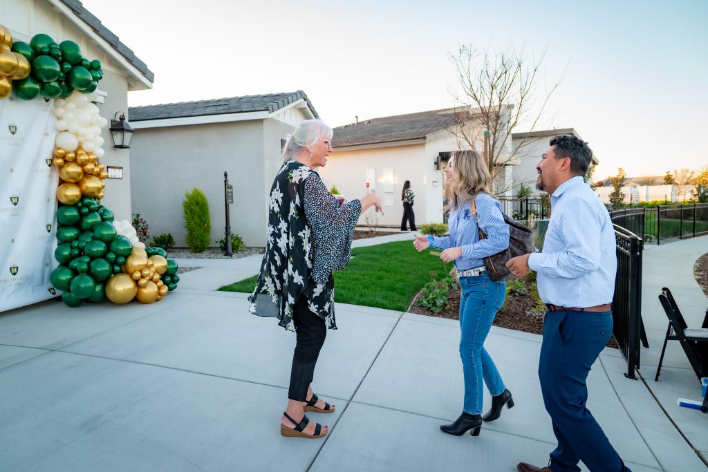 Three people greet each other outside a modern house decorated with green, gold, and white balloons during daylight, possibly at a celebration or open house event.