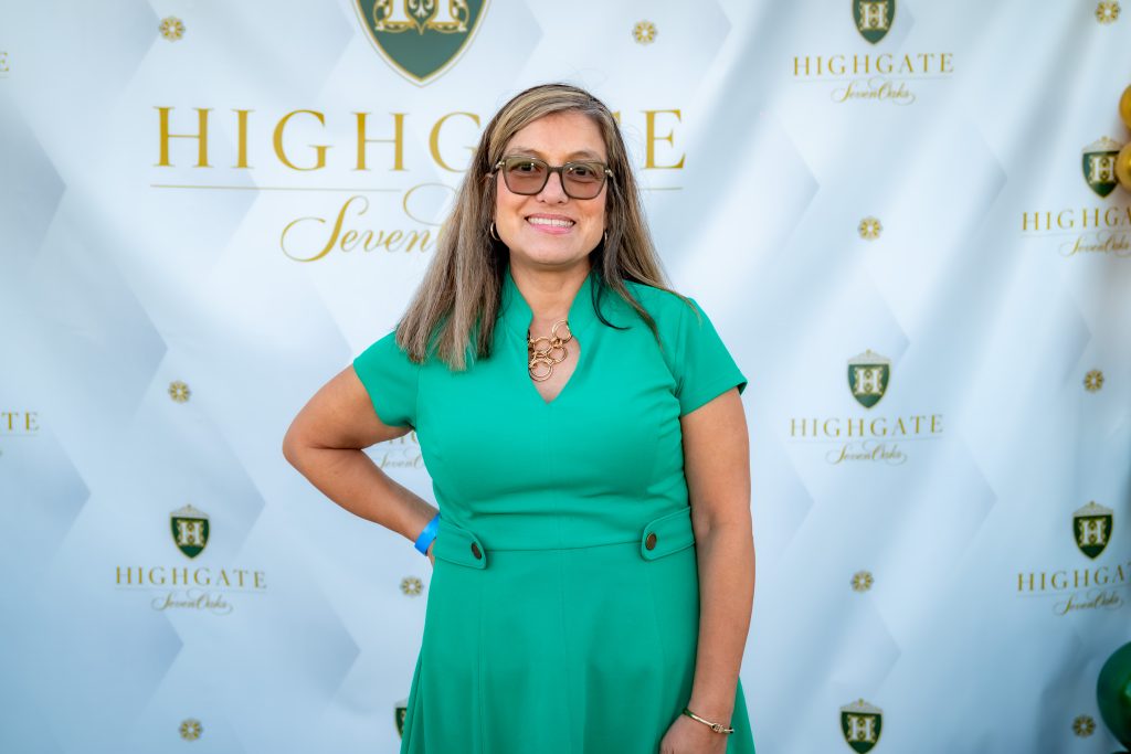 A woman in a green dress stands smiling in front of a white backdrop with gold "Highgate" and shield logos. She has glasses, straight hair, and wears a gold necklace.