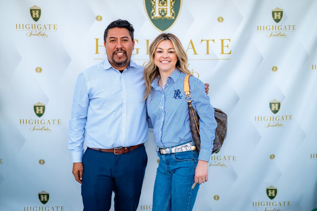 A man and a woman stand smiling together in front of a step-and-repeat banner with the "Highgate Jewel Oaks" logo. Both are dressed in blue outfits, and the woman carries a brown shoulder bag.