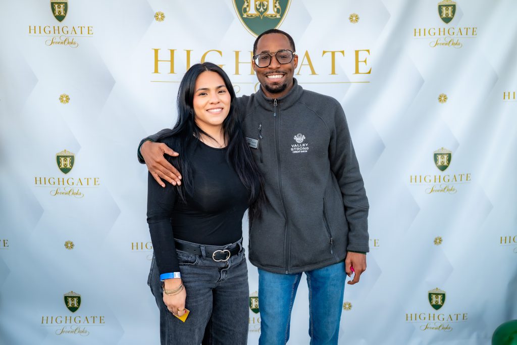 A woman and a man stand smiling together in front of a step-and-repeat banner with the Highgate logo and gold text. The man has his arm around the woman's shoulders. Both are casually dressed.