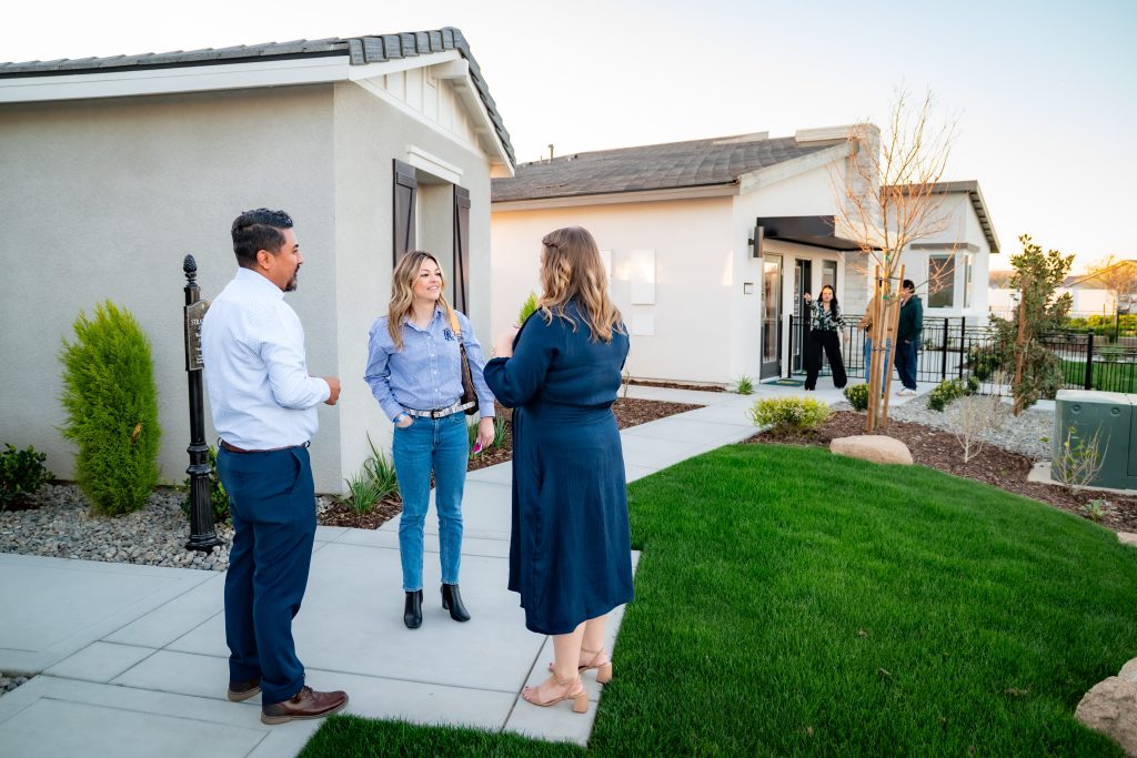 Three people converse on a sidewalk outside a modern house, while two others stand near the entrance in the background. The scene is set on a green lawn with landscaping and clear skies.