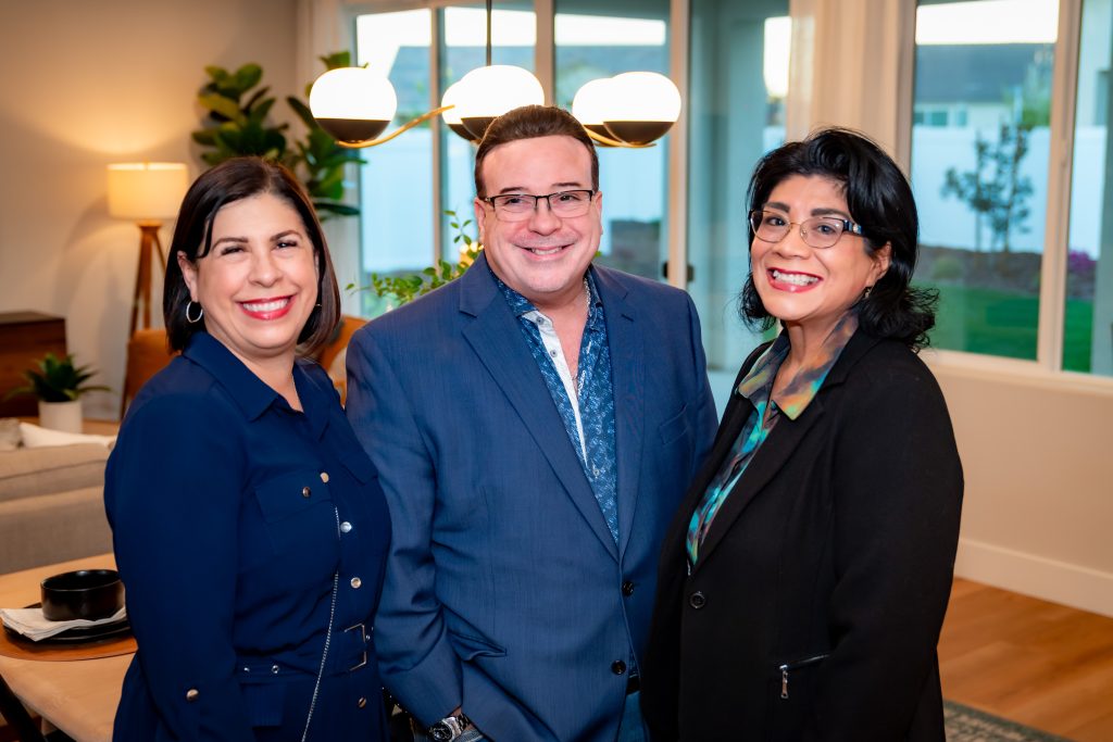 Three adults stand smiling indoors, dressed in business attire. Two women in dark jackets are on either side of a man in a blue suit. They are in a well-lit, modern living room with large windows in the background.