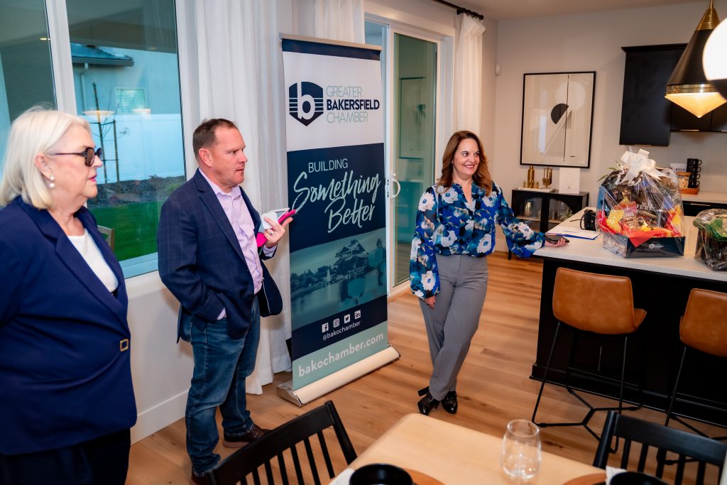 Three people stand and talk in a modern kitchen near a "Greater Bakersfield Chamber" banner. One woman gestures toward a counter with a gift basket. A glass sits on the table in the foreground.