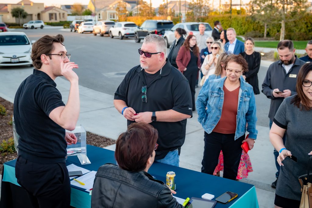 A group of people stands in line at an outdoor registration table on a neighborhood street, while two event staff assist them. The atmosphere is casual and social, with houses visible in the background.
