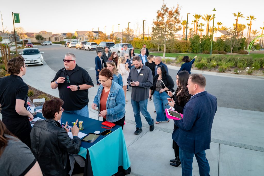 A group of people stand in line outdoors at a check-in table for an event, interacting with staff and using phones, with houses and greenery visible in the background.