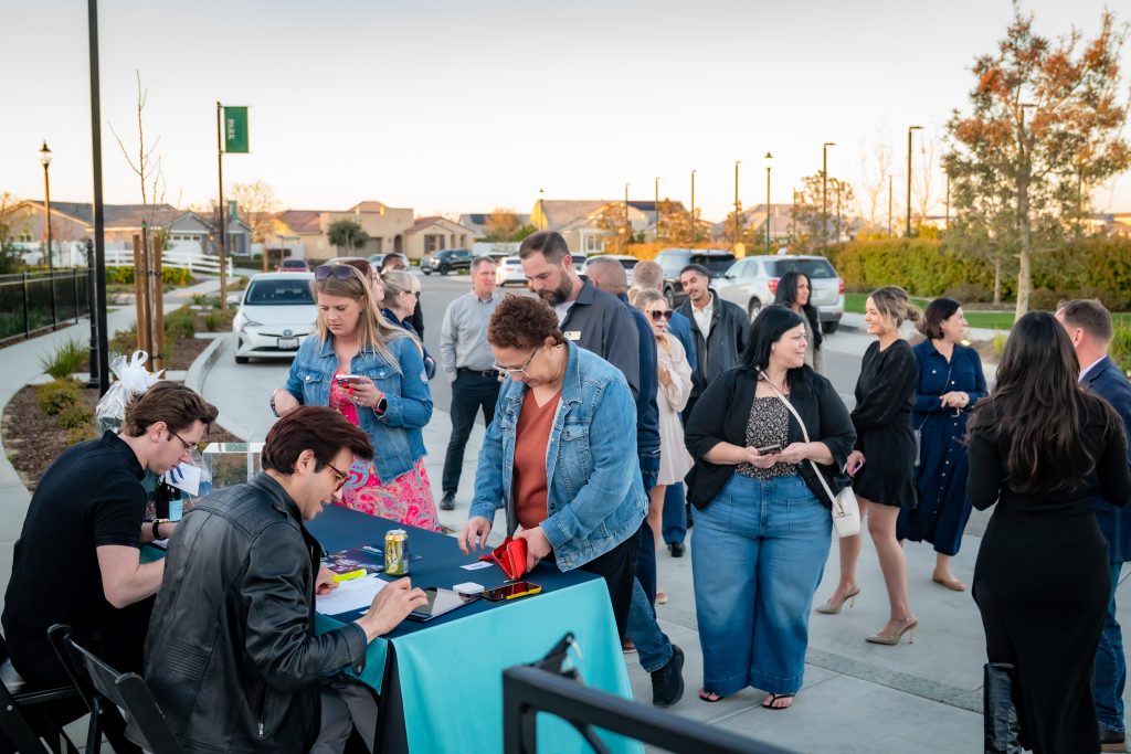 A group of people gathers outdoors near a table where two men are checking in guests. Some people are holding cards or chatting, and cars are parked in the background on a sunny day.