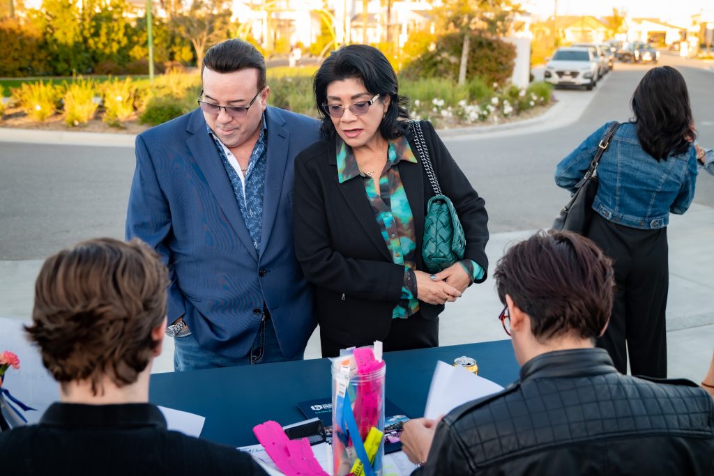 A man and woman stand together at an outdoor check-in table, speaking with two people seated across from them. The background shows a sunny street and greenery.
