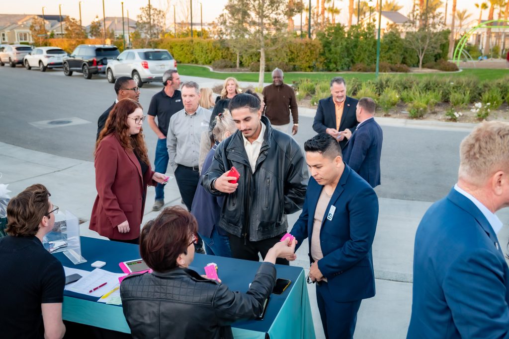 A group of people stand in line at an outdoor registration table, receiving name tags and colorful items from staff. Cars are parked along the street and trees are in the background.