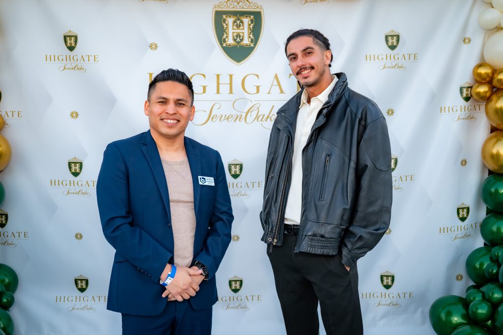 Two men pose in front of a Highgate Seven Oaks backdrop decorated with green, gold, and white balloons. One wears a blue suit and the other a black leather jacket and light shirt. Both are smiling and standing side by side.