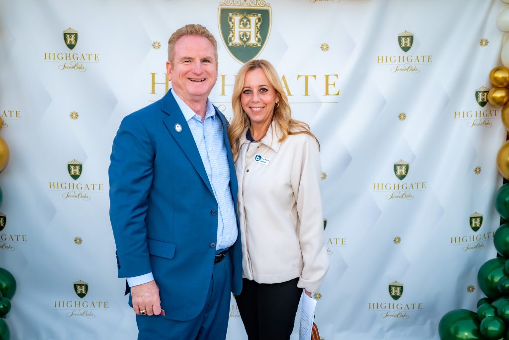 A man in a blue suit and a woman in a light jacket stand smiling in front of a Highgate-branded backdrop decorated with gold, green, and white balloons.