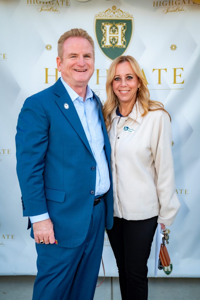 A man in a blue suit and a woman in a cream jacket stand smiling together in front of a step-and-repeat backdrop with a gold "Highgate" logo and text.