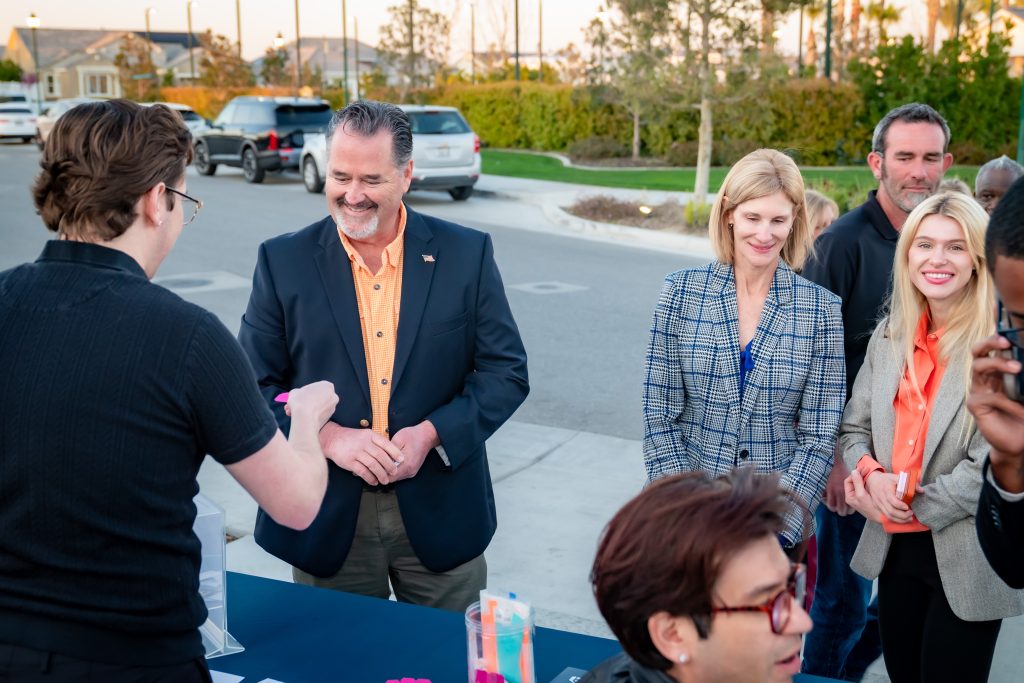 Several adults stand outdoors in a small group, smiling and interacting near a table. One man faces them, appearing to check people in or hand out items. Cars and greenery are visible in the background.