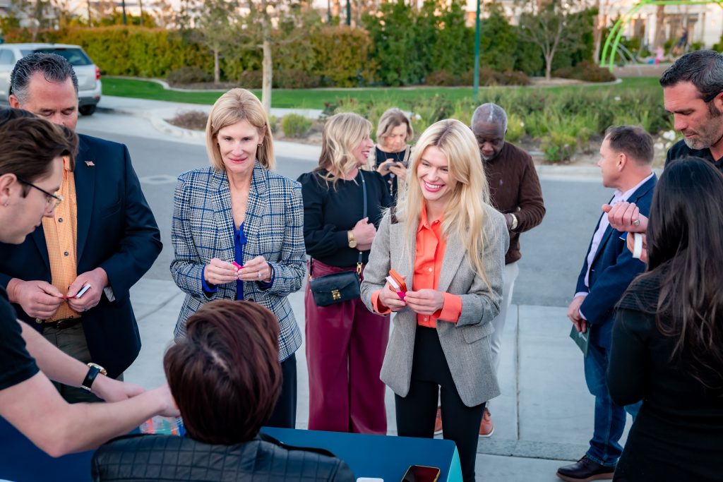 A group of people stand outdoors near a table, engaging in conversation and holding name badges. Some are smiling, and others are waiting in line, with greenery and parked cars in the background.