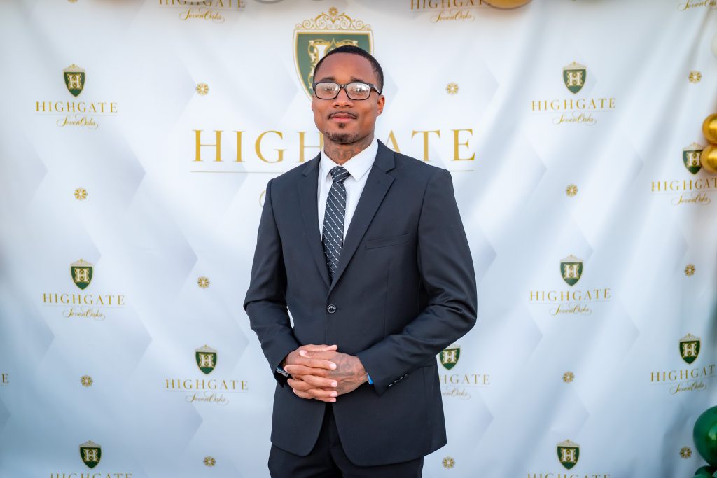 A man in a black suit, striped tie, and glasses stands in front of a white backdrop with "Highgate" and a green and gold crest printed on it. He is facing the camera and has his hands clasped.