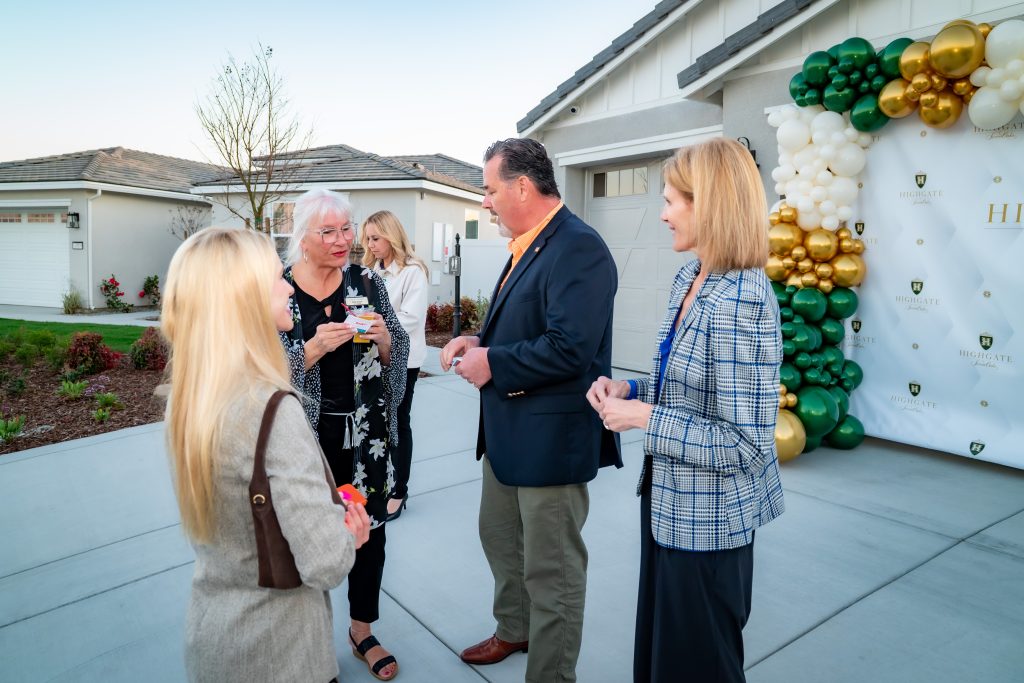 Five adults stand outside near a decorated house, having a conversation. There are green, white, and gold balloons and a branded backdrop in the background, suggesting a celebratory or formal event.