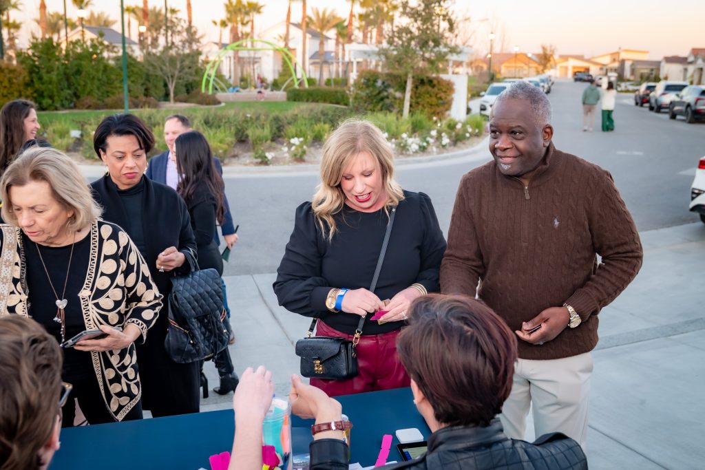 A group of people stands outdoors at a registration table, interacting with the staff. There are houses, palm trees, and a park in the background, suggesting a community event.